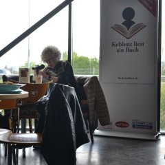 Eine Frau sitzt an einem Tisch und liest aus einem Buch vor.