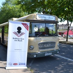 Ein Bus mit der Aufschrift "Oldie-Bus" steht neben einem Roll-Up-Poster mit dem Logo von Koblenz liest ein Buch.